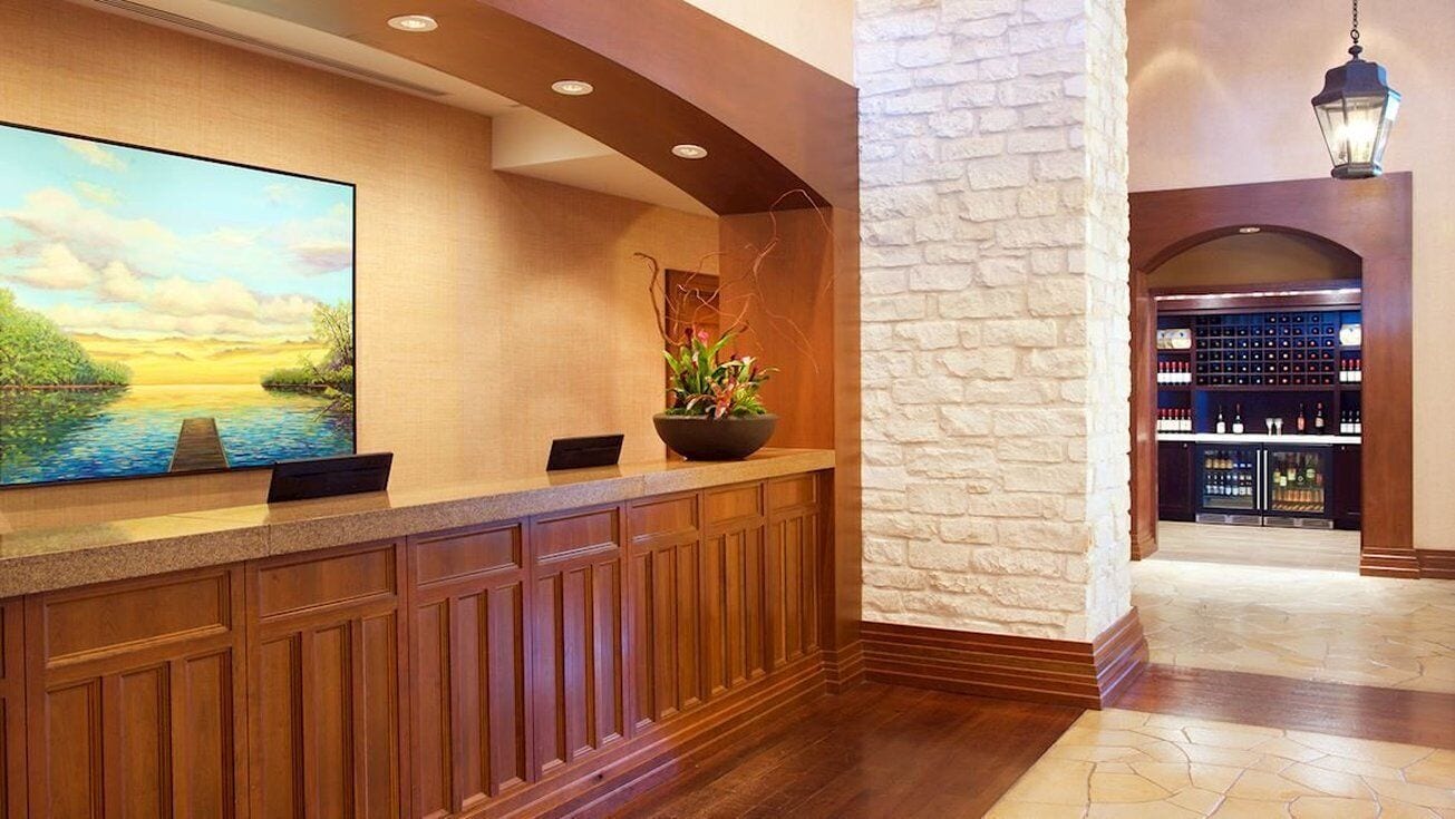 A reception area with a wooden counter, a large painting of a landscape, a stone wall, and a bar with shelves of bottles.