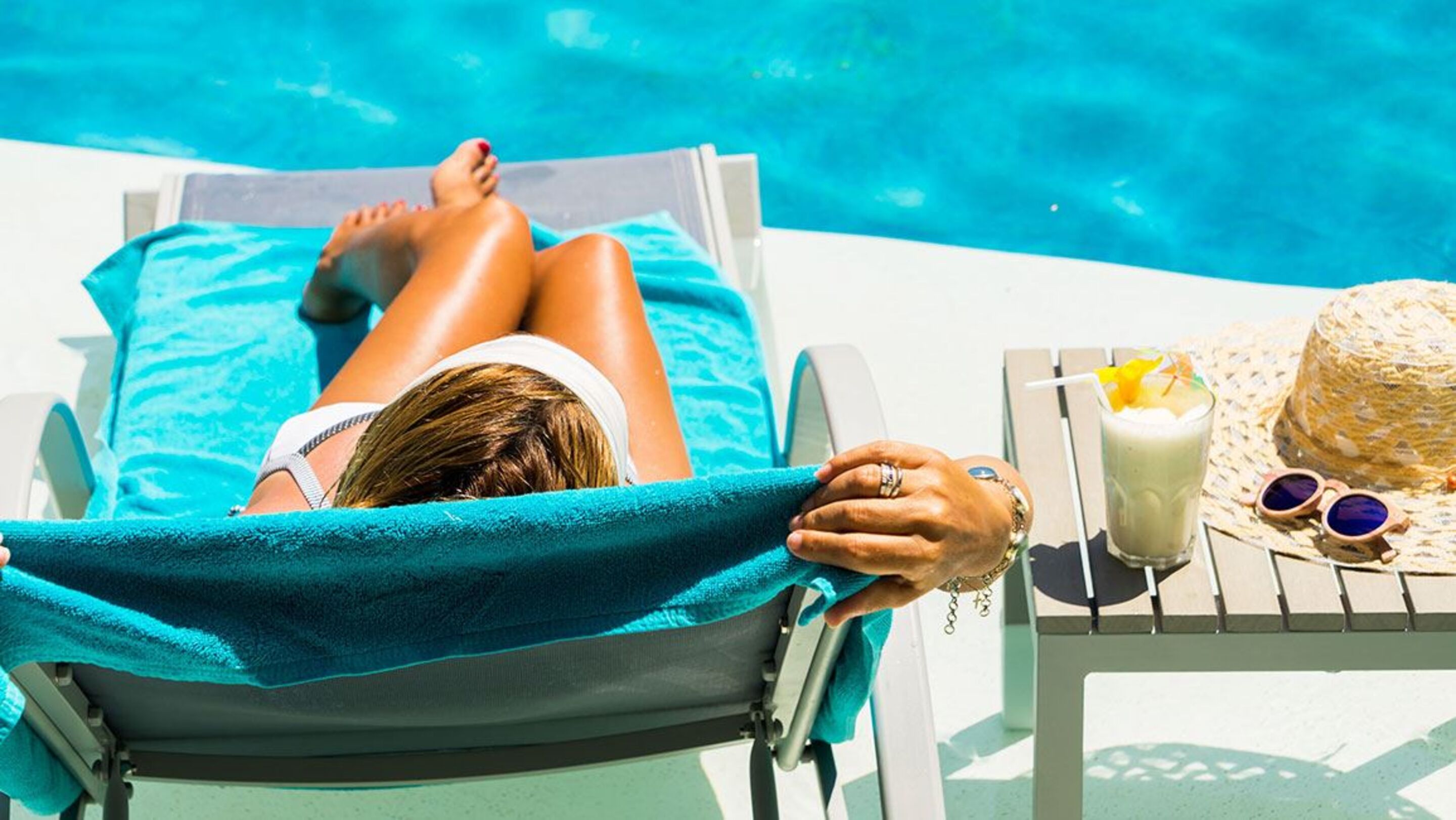 A person relaxing on a sun lounger by a pool with a drink and a straw hat on a table beside them.