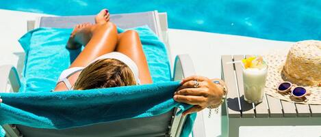 A person relaxing on a sun lounger by a pool with a drink and a straw hat on a table beside them.