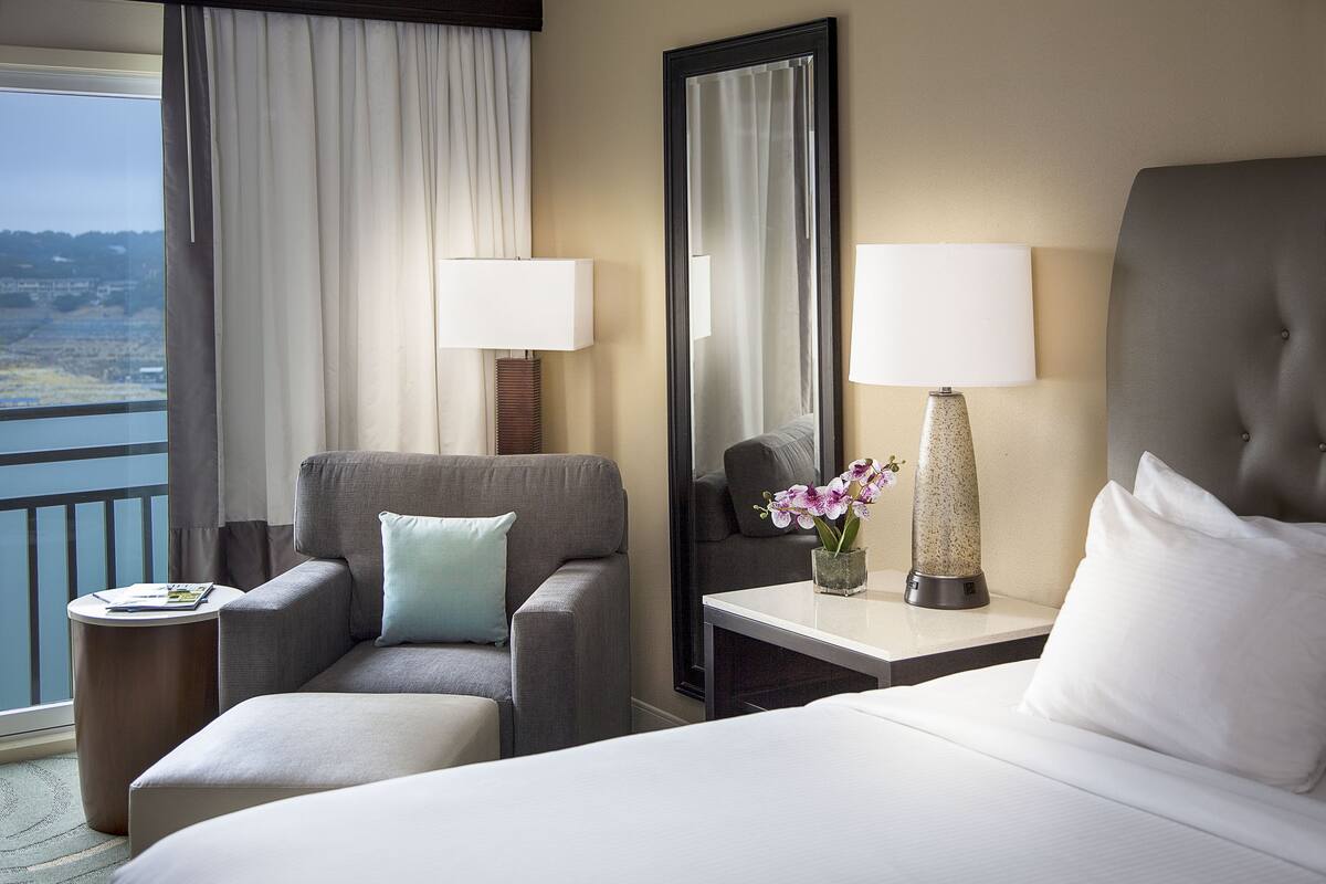 A hotel room with a bed, a sofa, a small table with a lamp, a mirror, and a view of the outdoors.
