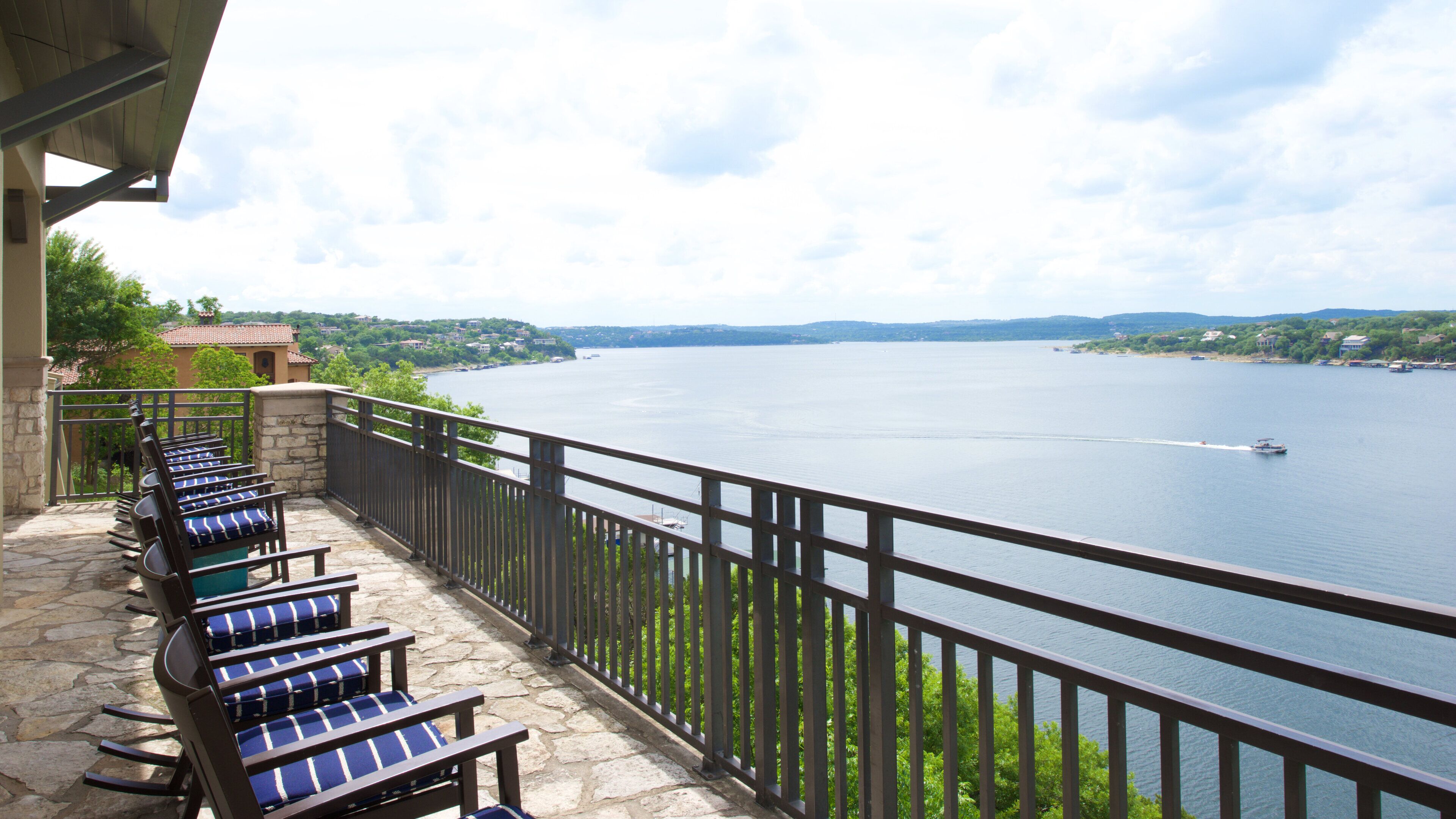 A balcony with stone flooring and metal railings, overlooking a calm lake with boats and houses in the distance.