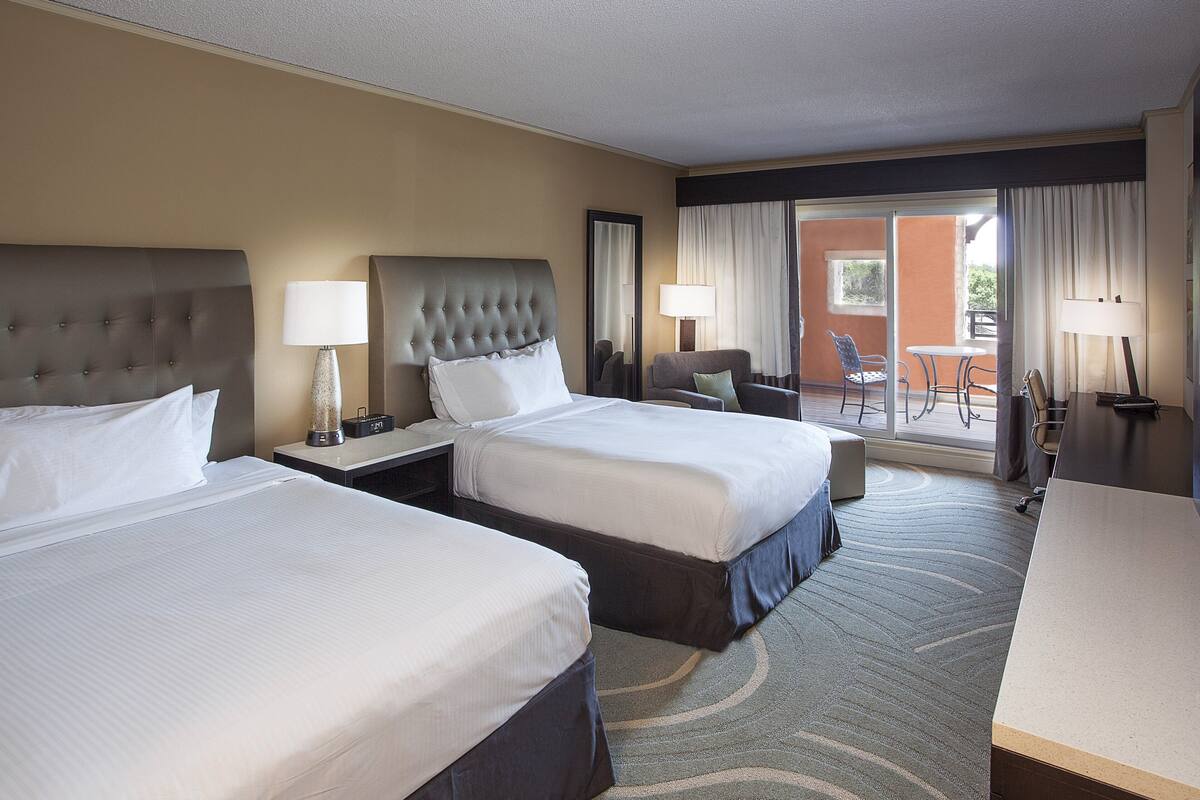 A hotel room with two beds, a desk, a chair, and a balcony with a table and chairs.