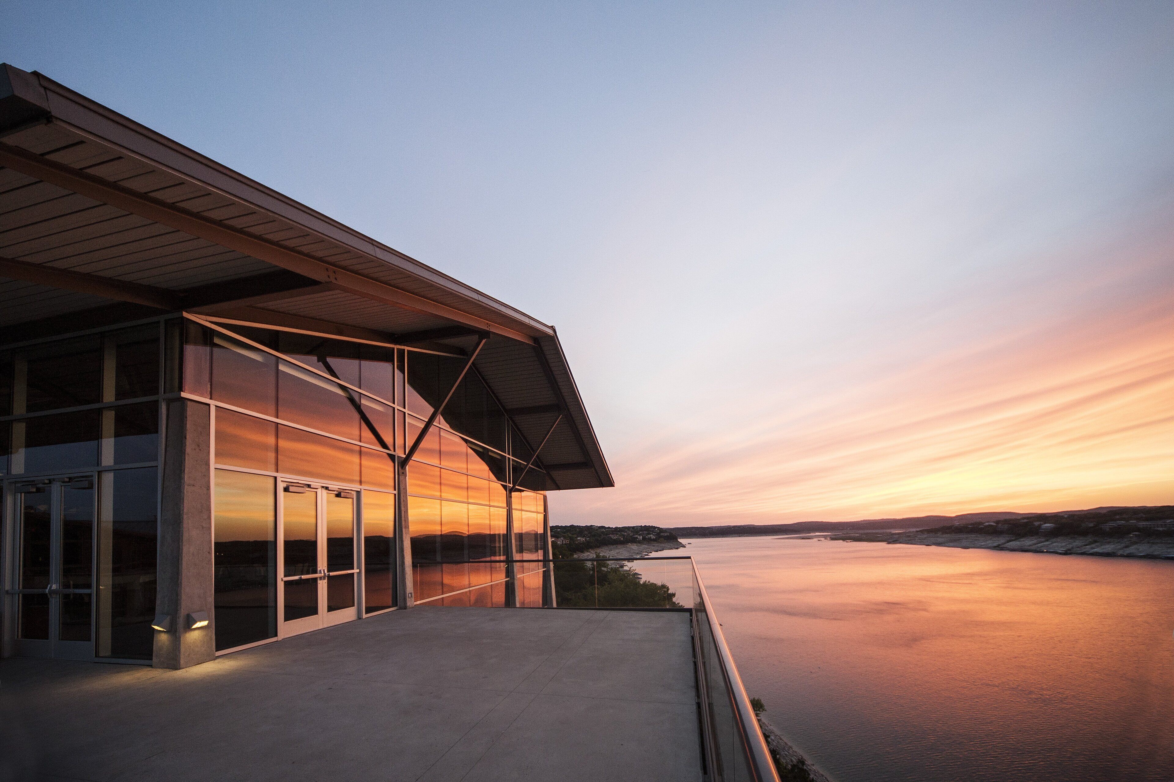 A modern building with large glass windows and a flat roof overlooking a calm body of water during sunset.