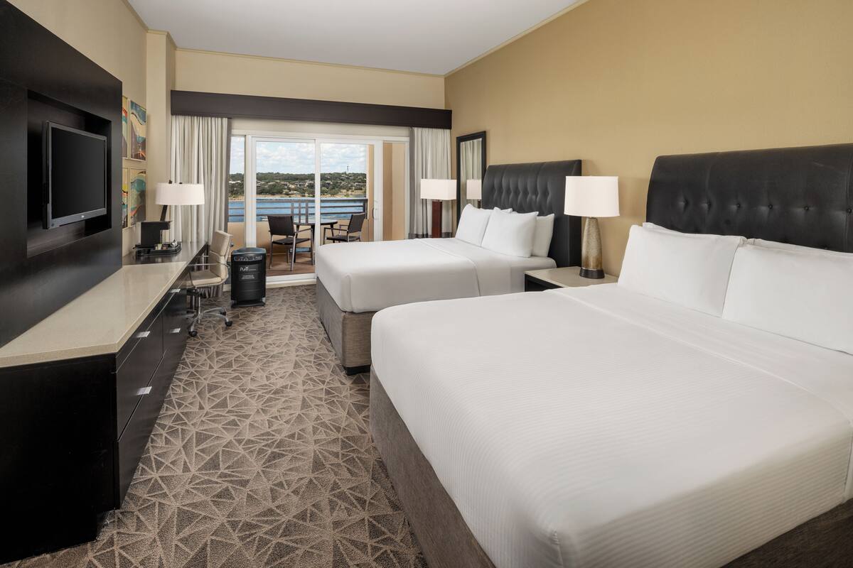 A hotel room with two beds, a desk, and a TV. There is a balcony with a view of the sea.