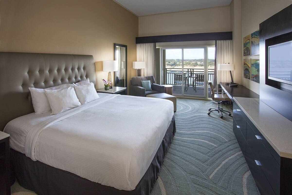 A hotel room with a large bed, a desk, a chair, and a balcony with a view.