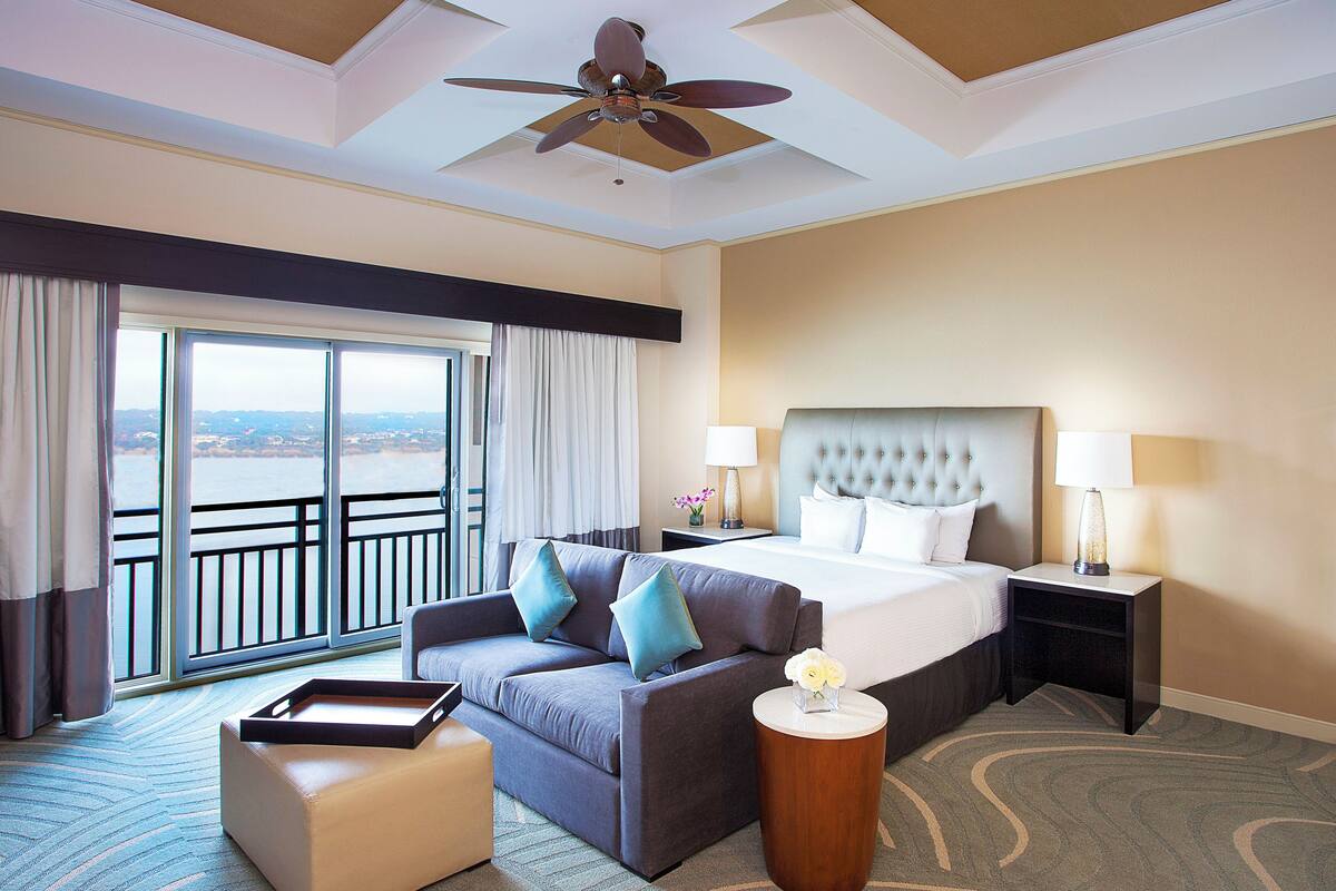 A hotel room with a bed, a sofa, a coffee table, a side table with a lamp, and a view of the outside.