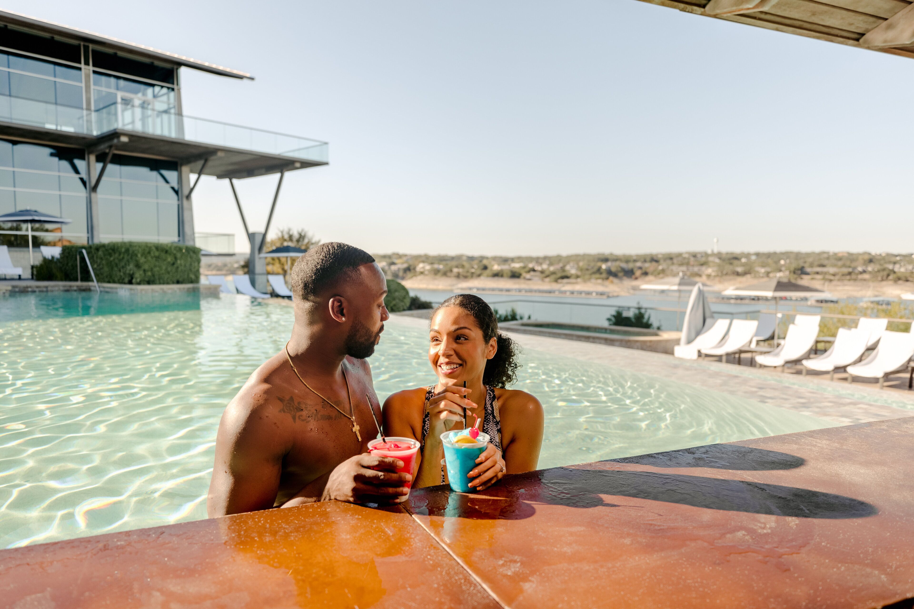 Una pareja junto a una piscina, con bebidas, un edificio moderno y sillones de descanso al fondo.
