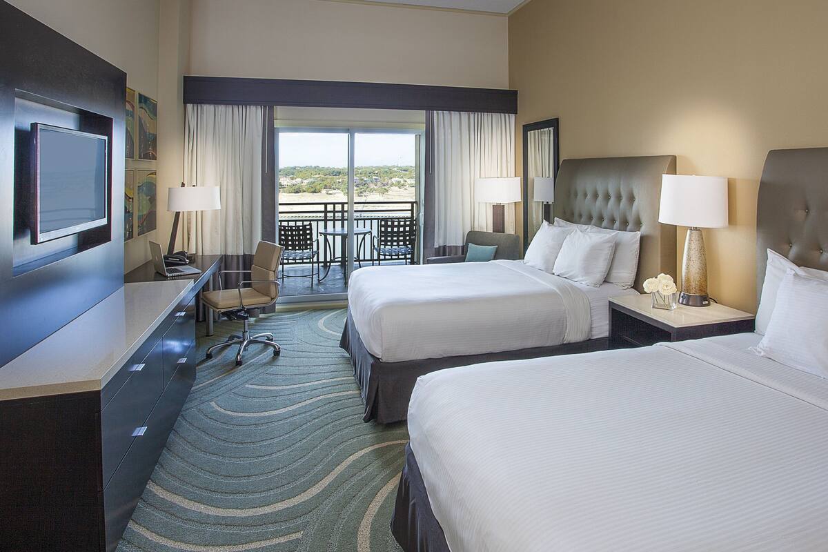 A hotel room with two beds, a desk, a chair, a TV, and a balcony with a view.