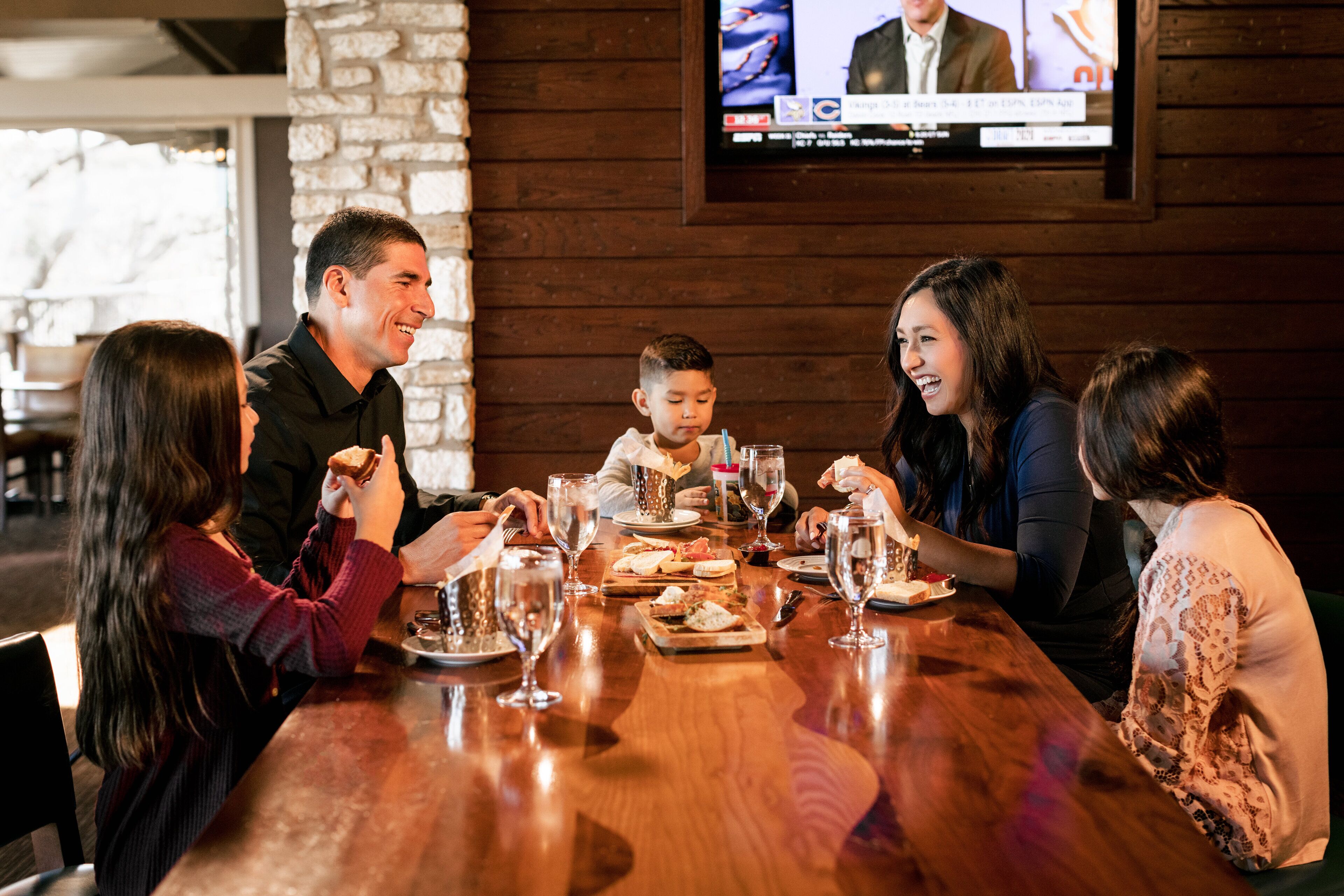 A group of people dining at a wooden table with a television mounted on the wall displaying a news broadcast.