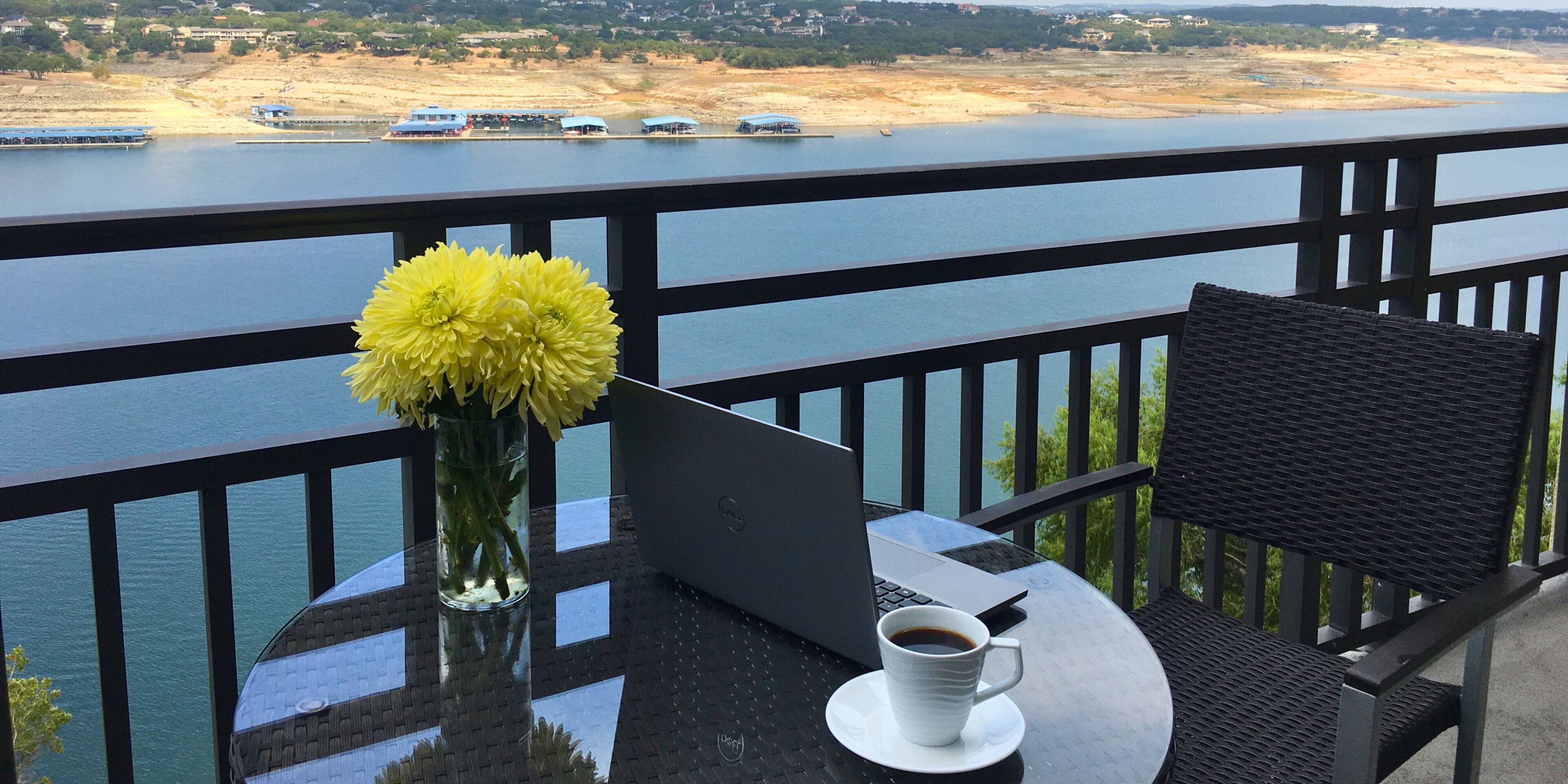 A balcony with a laptop, a cup of coffee, and a vase of yellow flowers.