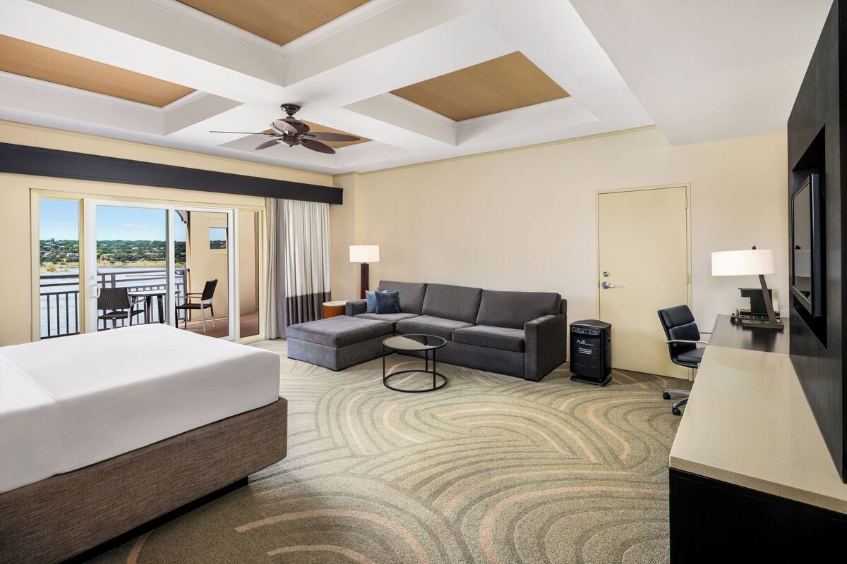 A hotel room with a bed, a sofa, a desk, a chair, a TV, a ceiling fan, and a view of the outdoors.