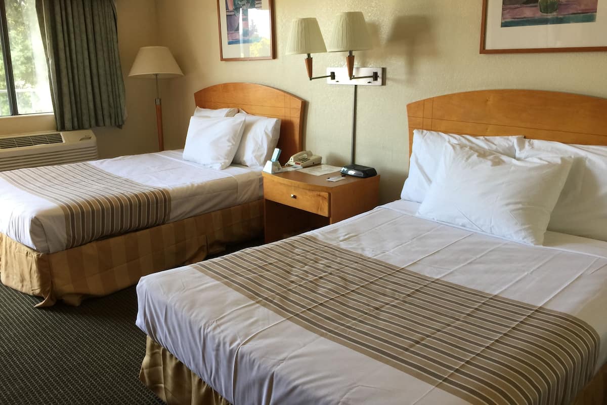 A hotel room with two beds, a bedside table with a lamp, a wall-mounted phone, and a window with curtains.