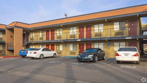 A motel with a row of rooms, each with a red door and a window. There are cars parked in front of the motel. - Fresno Inn (Fresno)