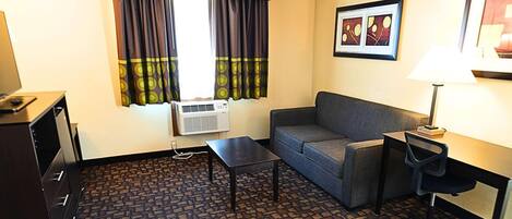 Habitación de hotel con sofá, una mesita, escritorio con silla, unidad de aire acondicionado y ventana con cortinas.