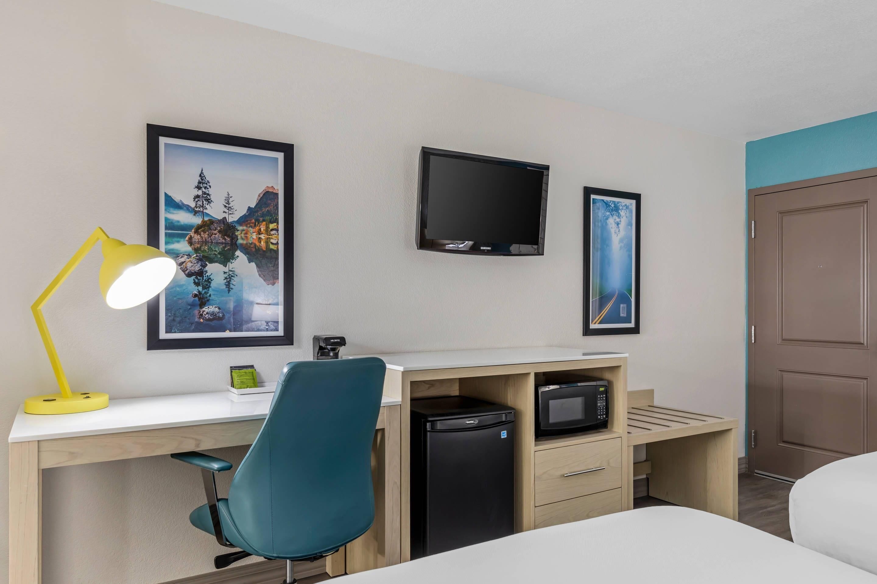 A hotel room with a desk, a chair, a television, a refrigerator, and a door.