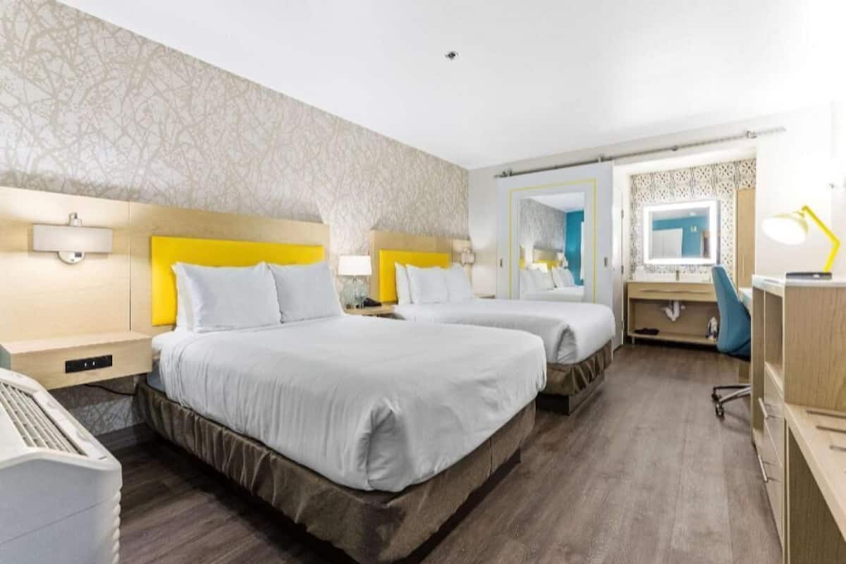 A hotel room with two beds, a desk, a chair, a mirror, and a lamp.