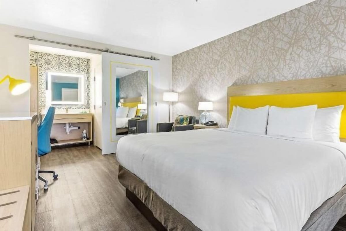 A hotel room with a large bed, bedside tables, a desk with a chair, a mirror, and a patterned wall.