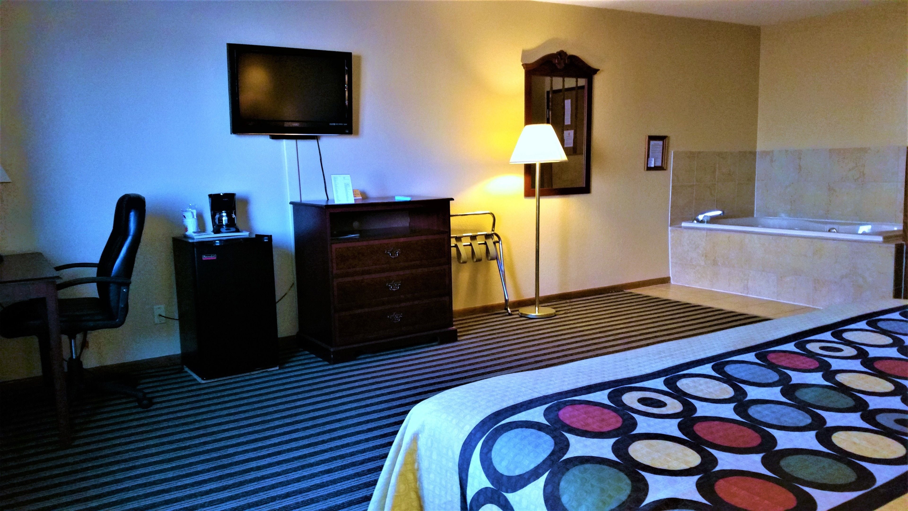Una habitación de hotel con cama, escritorio con silla, televisor, cómoda, lámpara, toallero y bañera.