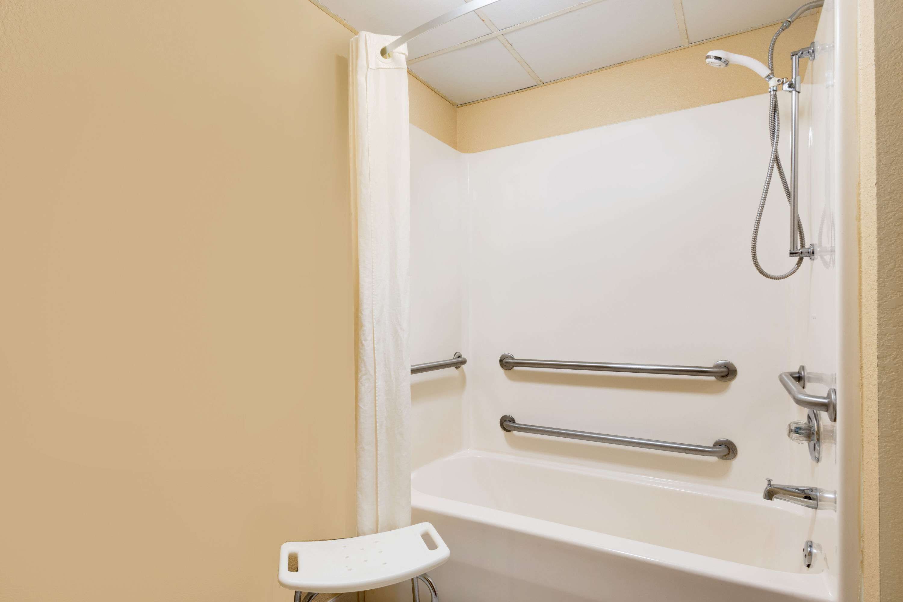 Une salle de bain avec une baignoire blanche, des barres d’appui, une pomme de douche et un siège de douche blanc.