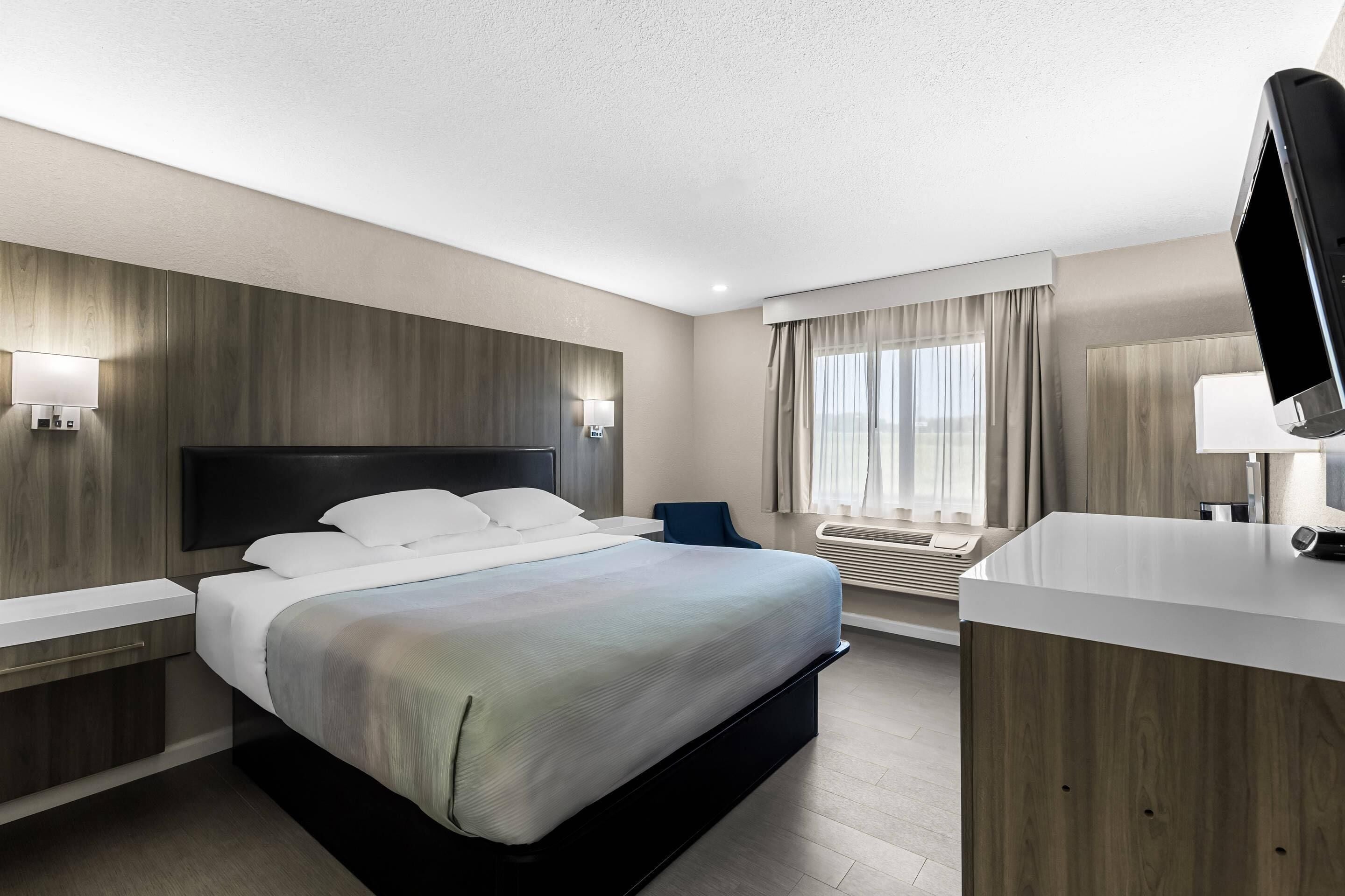 A hotel room with a bed, bedside tables, a desk, a chair, a TV, a window with curtains, and a wall-mounted air conditioning unit.