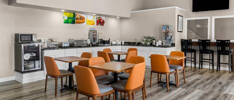 Cocina moderna con microondas, refrigerador y cafetera. Hay un comedor con sillas naranjas y una mesa.