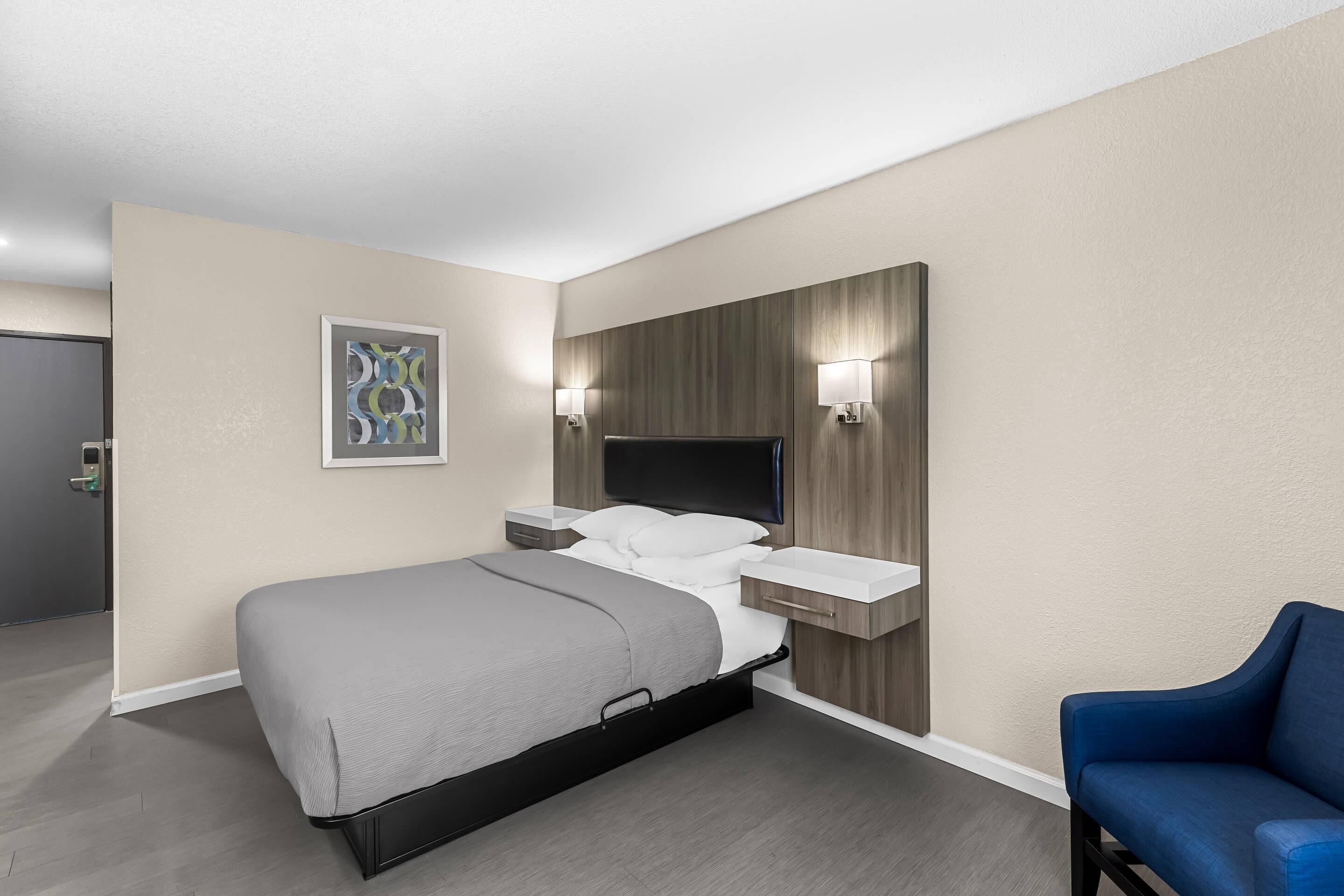Habitación de hotel con cama, mesitas de noche, televisión, estante en la pared, una silla y un cuadro.