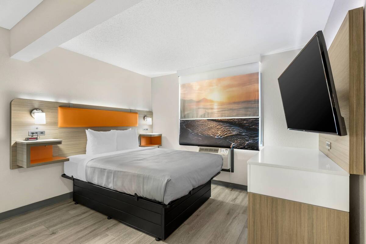 A modern hotel room with a bed, bedside tables, a flat-screen TV, and a large window with a sunset view.