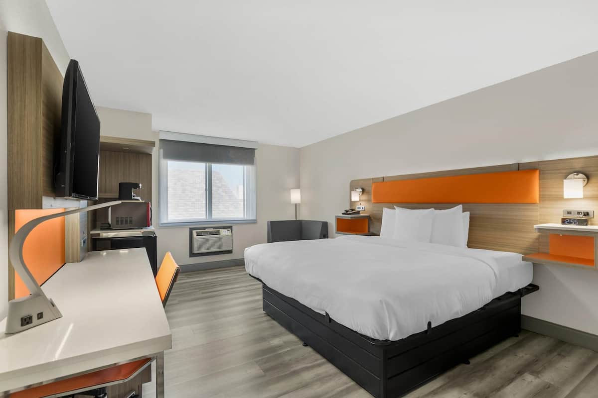 A hotel room with a bed, desk, chair, TV, bedside table, lamp, and window.