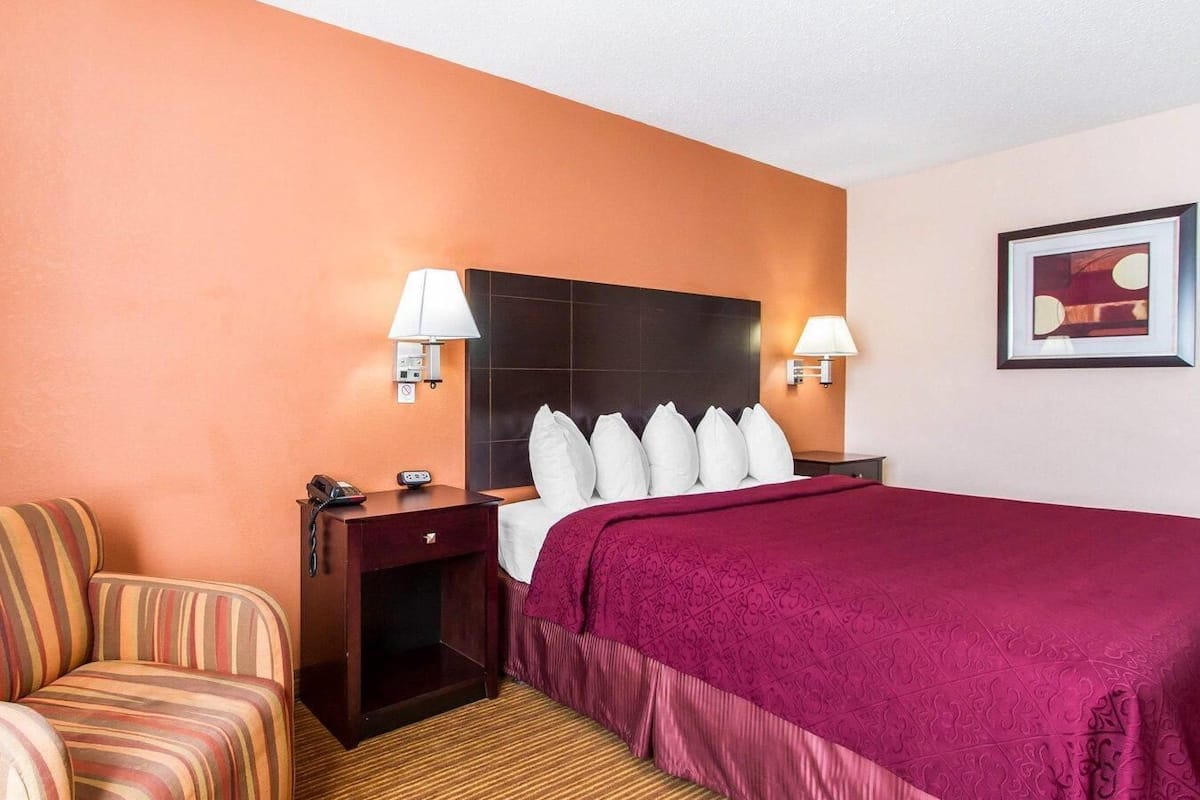 A hotel room with a bed, two bedside lamps, a chair, a nightstand, and a framed picture on the wall.