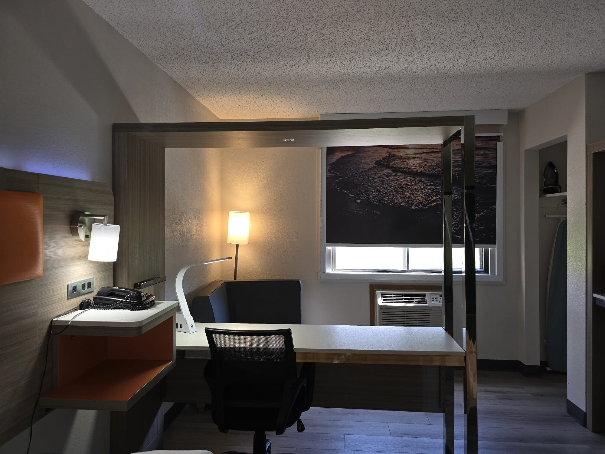 A hotel room with a desk, chair, lamp, and a large window with a view.