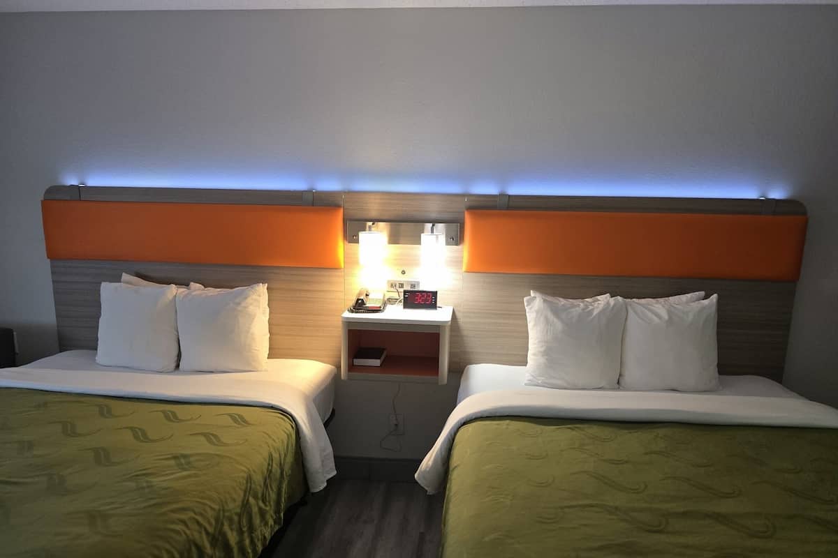 A hotel room with two beds, each with white bedding and green headboards. There is a nightstand with a lamp and a digital clock between the beds.