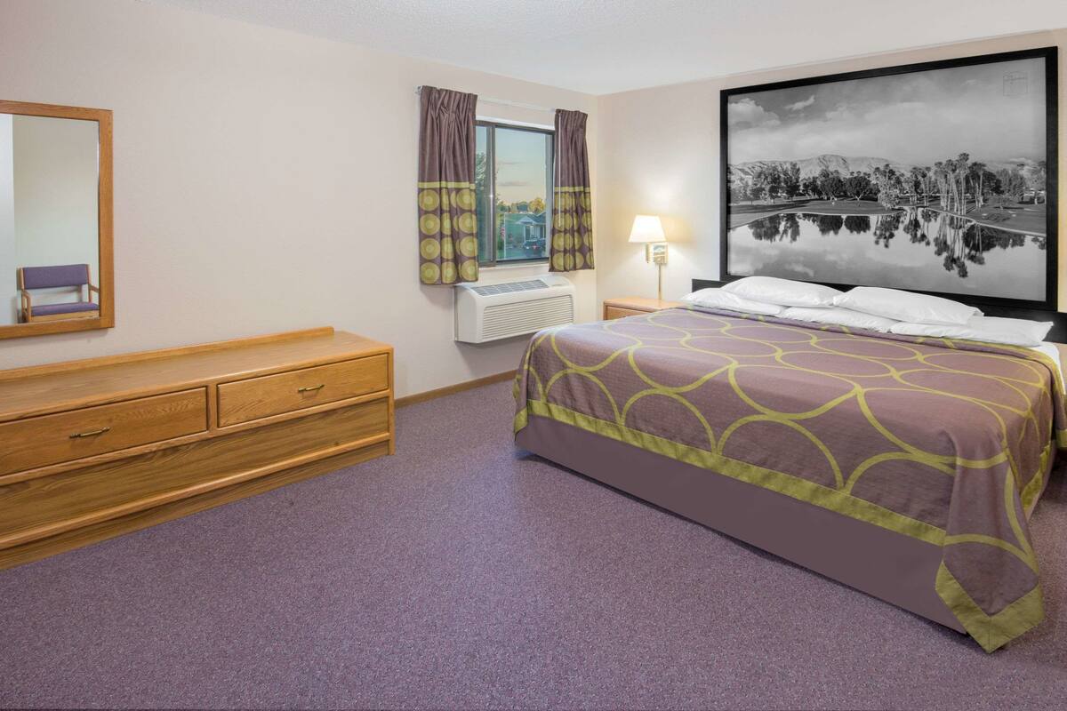A hotel room with a bed, a dresser, a window with curtains, a wall-mounted air conditioner, and a large framed picture on the wall.