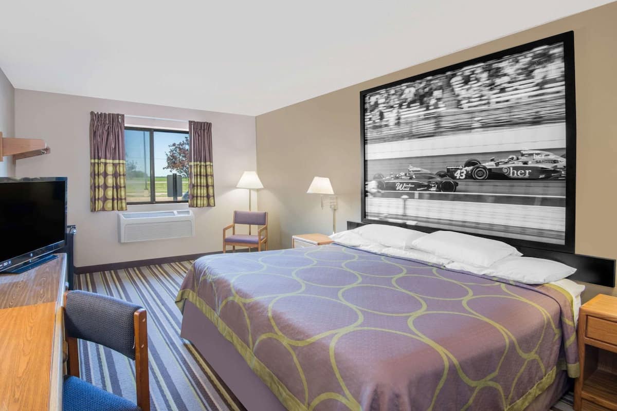 A hotel room with a bed, a desk with a TV, a chair, a window with curtains, and a wall-mounted picture.