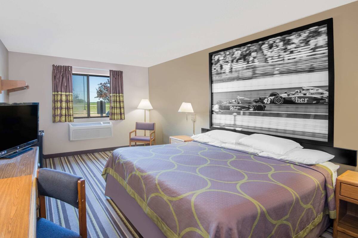 A hotel room with a bed, a desk with a TV, two lamps, a chair, and a large framed picture of a racing car on the wall.