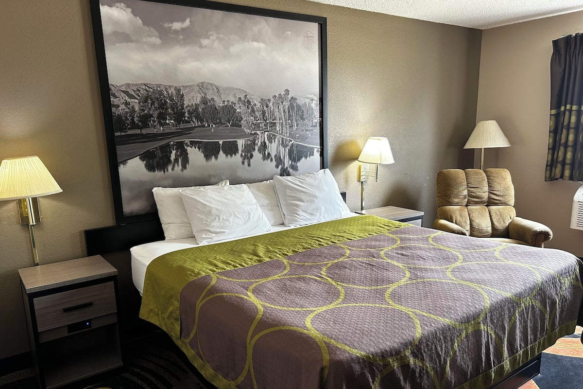 A hotel room with a bed, two bedside lamps, a chair, a nightstand, and a large framed picture on the wall.