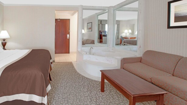 A hotel room with a bed, a sofa, a small table, a bathroom with a bathtub, and a mirror. - Baymont by Wyndham Jackson (Jackson)