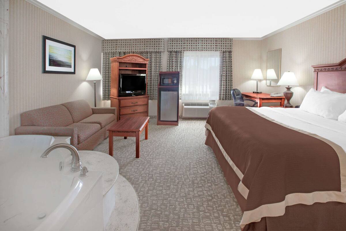A hotel room with a bed, a sofa, a desk, a television, a refrigerator, a lamp, and a bathroom with a bathtub and sink.