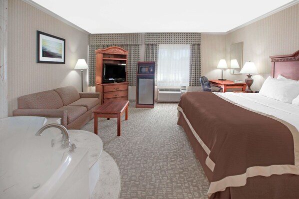 A hotel room with a bed, a sofa, a desk, a television, a refrigerator, a lamp, and a bathroom with a bathtub and sink. - Baymont by Wyndham Jackson (Jackson)