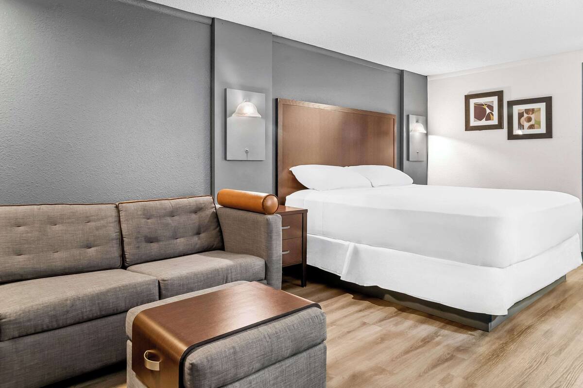 A hotel room with a grey sofa, a wooden side table, a bed with white bedding, and two wall-mounted light fixtures.