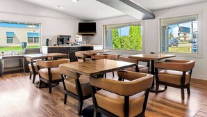 Een moderne eetruimte met houten tafels en lederen stoelen, een keuken met een flatscreen televisie, en grote ramen met uitzicht op groen en een gebouw.