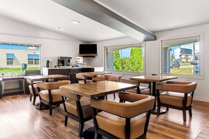 Un comedor moderno con mesas de madera y sillas de cuero, una zona de cocina con televisor de pantalla plana y ventanas amplias que ofrecen vistas a zonas verdes y un edificio exterior.