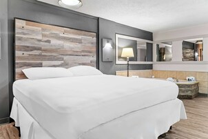Habitación de hotel con una cama grande, cabecera de madera, mesita de noche con lámpara, espejo y un área pequeña de baño.