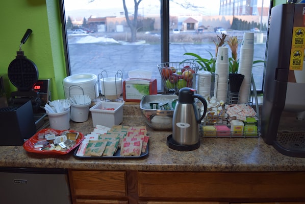 A kitchen counter with various items including a waffle maker, coffee maker, and food trays. - Super 8 by Wyndham Saginaw (Saginaw)