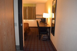 A hotel room with a bed, a desk with a computer, a chair, a trash bin, and a mirror on the wall.
