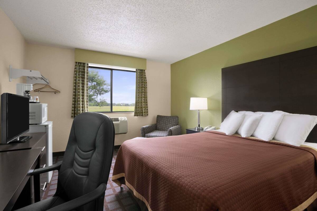Habitación de hotel con cama, escritorio con computadora, silla, televisor y ventana con cortinas.