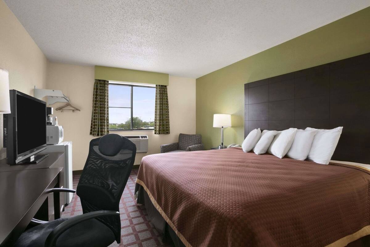 Una habitación de hotel con cama, escritorio con televisor, silla, lámpara y ventana con cortinas.
