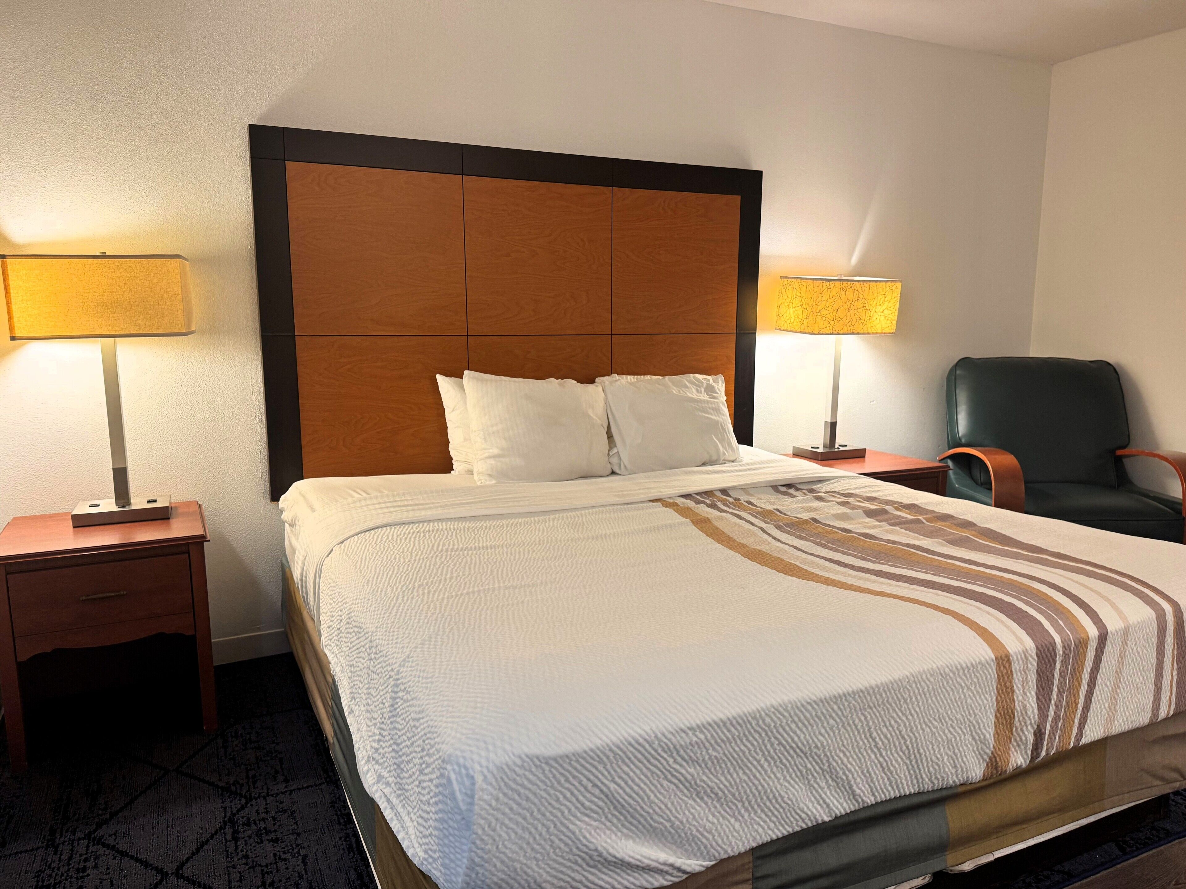 Une chambre d’hôtel avec un grand lit, deux tables de chevet avec des lampes, un fauteuil vert et une tête de lit en bois.