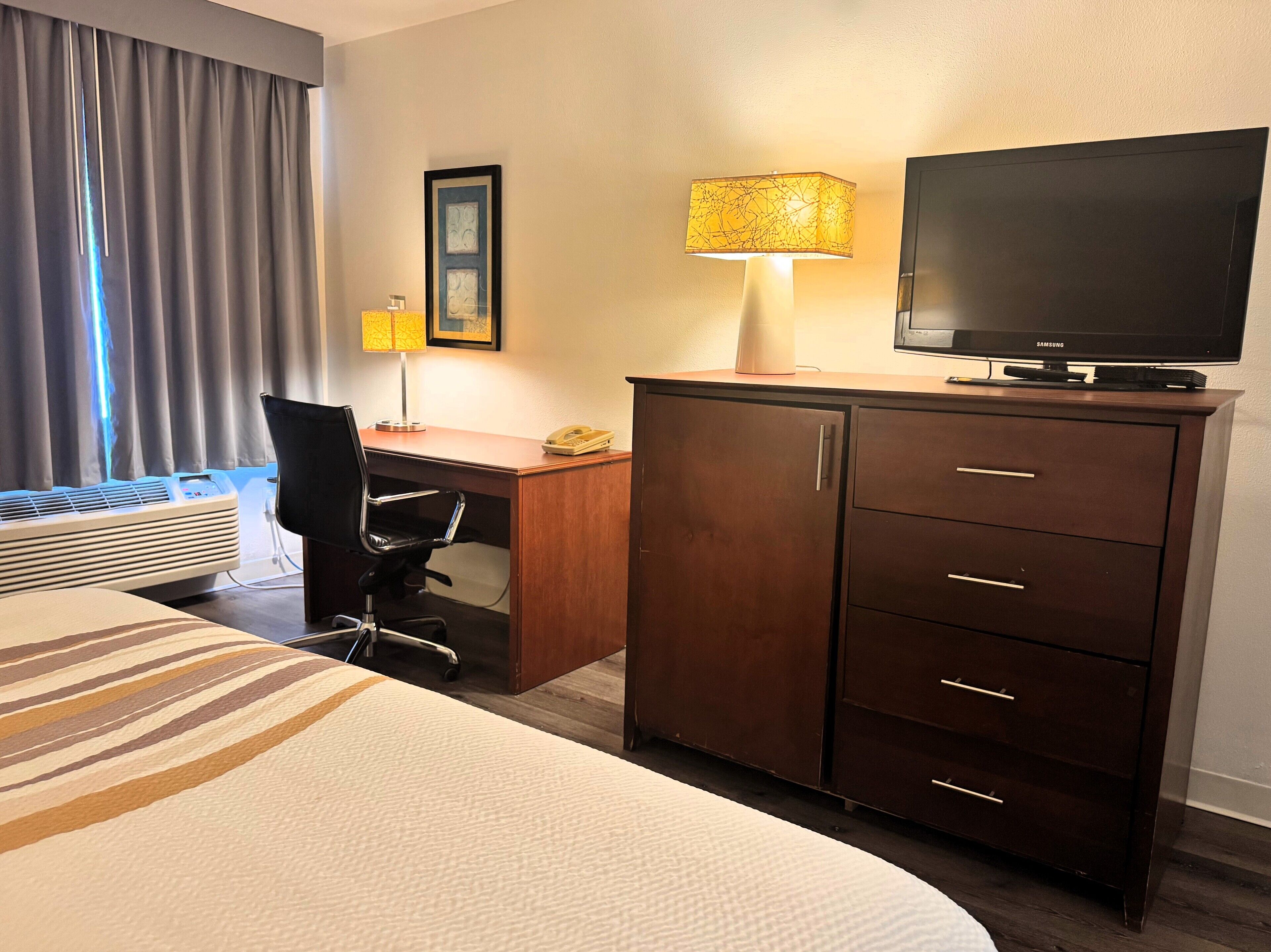 A hotel room with a bed, a desk, a television, a lamp, a chair, and a wardrobe.