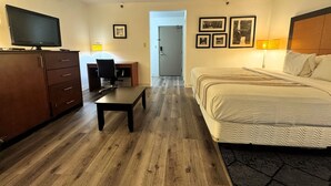 Quarto de hotel com piso de madeira, uma cama, uma escrivaninha com cadeira, uma televisão e uma porta que dá para outro cômodo.