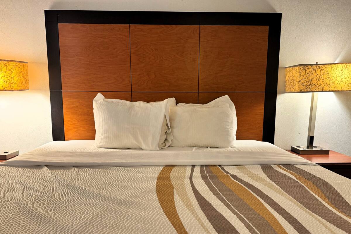 A hotel room with a large bed, two white pillows, a wooden headboard, and two bedside lamps.